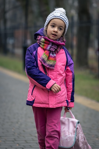 Free Photo little girl with a backpack in a jacket and a hat near the school