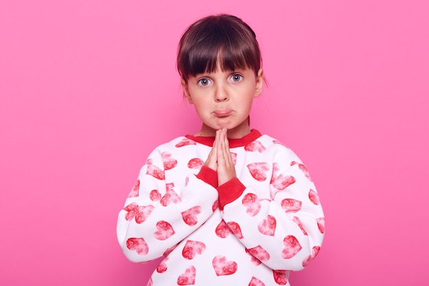 little girl with dark hair looks at camera with a pleading expression on her face, keeping palms together, praying, wearing a sweater, isolated over pink wall.