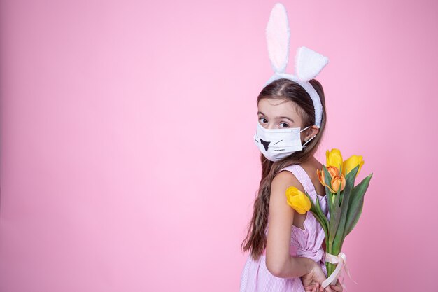 Little girl with easter bunny ears and wearing a medical face mask holds a bouquet of tulips in her hands on a pink