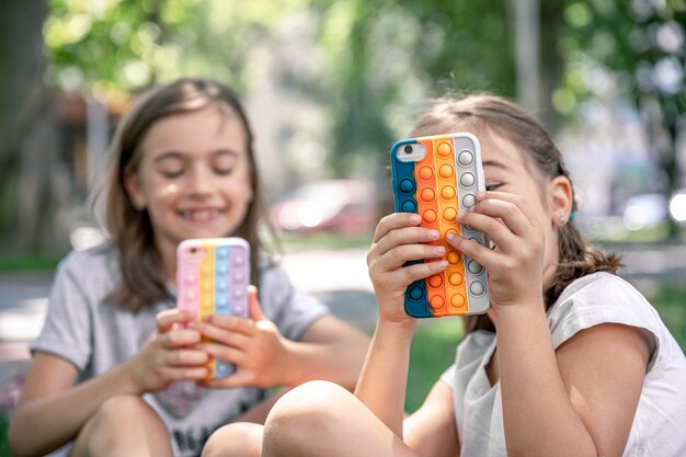 Little girls outdoors with phones in a case with pimples pop it, a trendy anti stress toy.