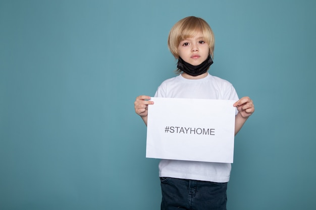 Free photo little kid in black protective mask with white t-shirt and blue jeans with stay home hashtag against covid on blue desk