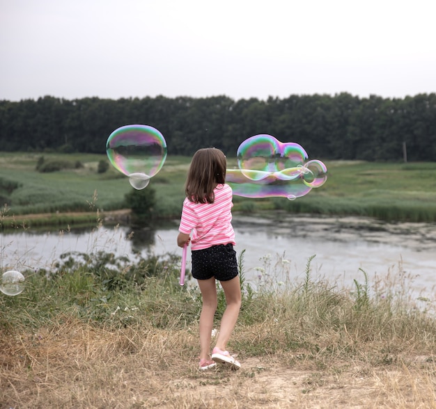 Little kid girl launches huge soap bubbles in the background beautiful nature, back view.