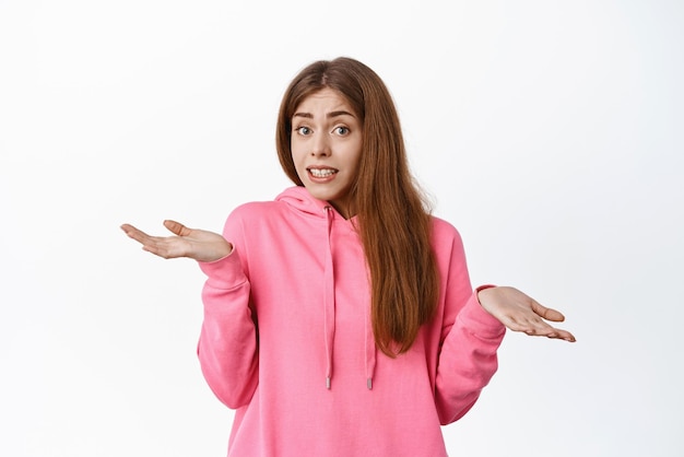 Little mistake oops Cute girl shrugging shoulders and look guilty say sorry dont know cant understand something standing indecisive over white background