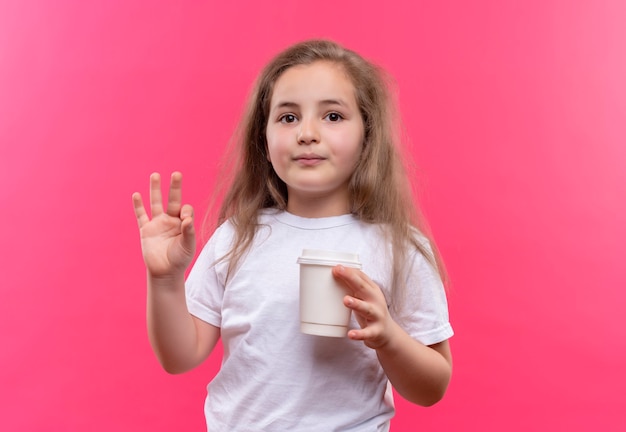 Free photo little school girl wearing white t-shirt holding cup of coffee showing okey gesture on isolated pink wall