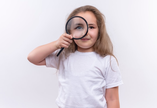 little school girl wearing white t-shirt looking through magnifier on isolated white wall