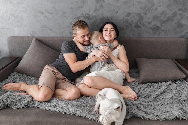 Free photo little son hugging her parents sitting on sofa with white dog