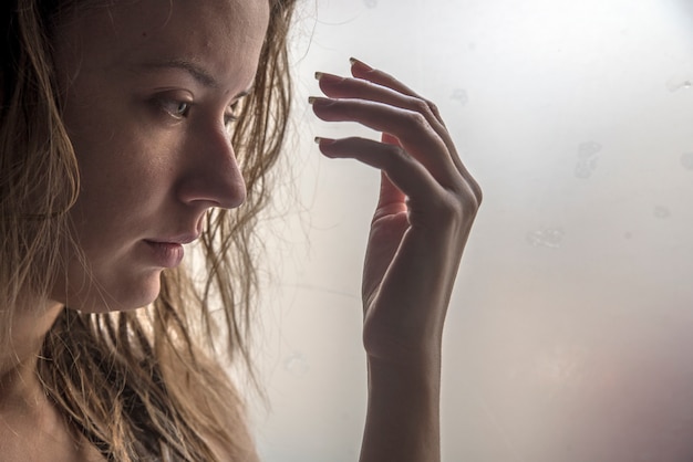 Free Photo lonely girl near window thinking about something. sad woman looking the rain falling through a window at home