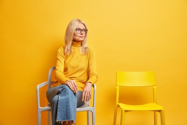 Free photo lonely middle aged woman sits near empty chair needs communication looks thoughtfully away dressed in casual turtleneck and jeans