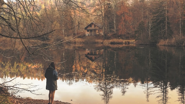 Free photo lonely woman standing near the lake with the reflection of the isolated wooden cabin visible