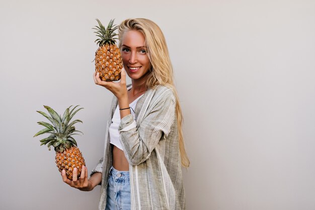 long-haired girl holding pineapple with charming smile. white lady posing with exotic fruits in studio.