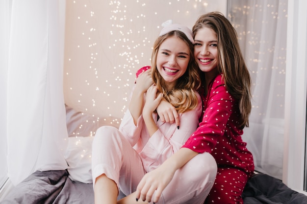 Long-haired girl posing emotionally near sister on bed. Indoor shot of spectacular female models in pajamas sitting in bedroom.