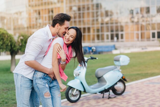 Long-haired smiling girl in ripped blue jeans having fun with boyfriend in city park. Outdoor portrait of funny loving couple fooling around on the street with scooter on background.