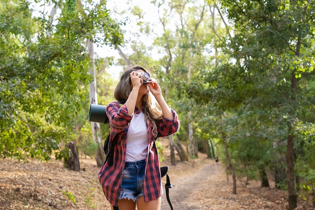 Free photo long-haired woman taking photo of nature and standing on road in forest. blonde caucasian lady holding camera and shooting landscape. backpacking tourism, adventure and summer vacation concept