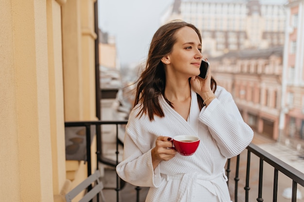 Long-haired young woman enjoying view of city on balcony. Girl in bathrobe drinks coffee and speaks on phone.