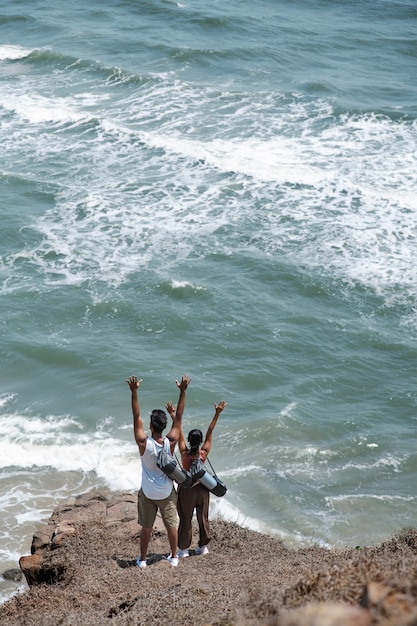 Free photo long shot couple with hands up at seaside