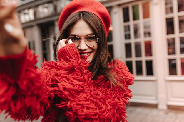 Longhaired brunette girl with white manicure and red lipstick makes selfie Shot of charming woman in glasses and red wool outfit against background of house