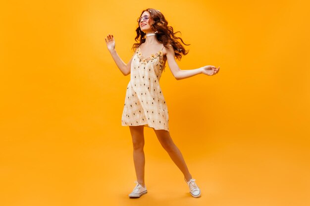 Longhaired curly lady in sunglasses and yellow sundress dancing on orange background