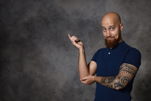 Look at that! Isolated funny bald bearded man with tattoo raising index fingre and pointing at left corner, expressing excitement or curiosity, raising eyebrows. Body language