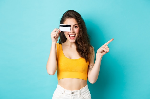 Free photo look there. stylish modern woman showing plastic credit card, smiling and pointing right, showing way to banner or logo, standing against blue background