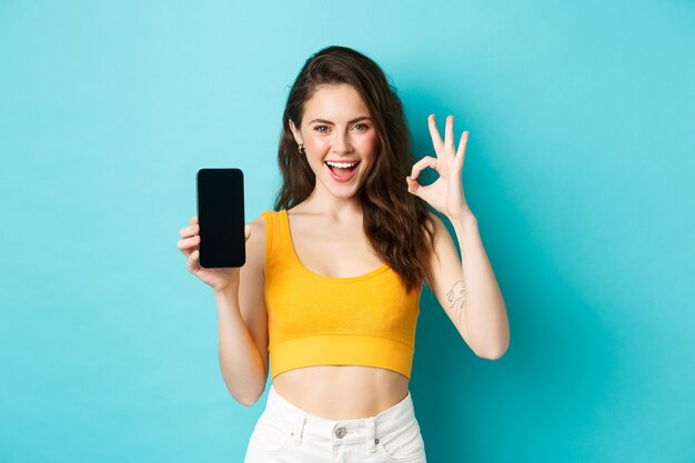 Look at this. Good-looking woman with sassy smile, winking and showing okay sign with empty smartphone screen, recommending app, standing over blue background.