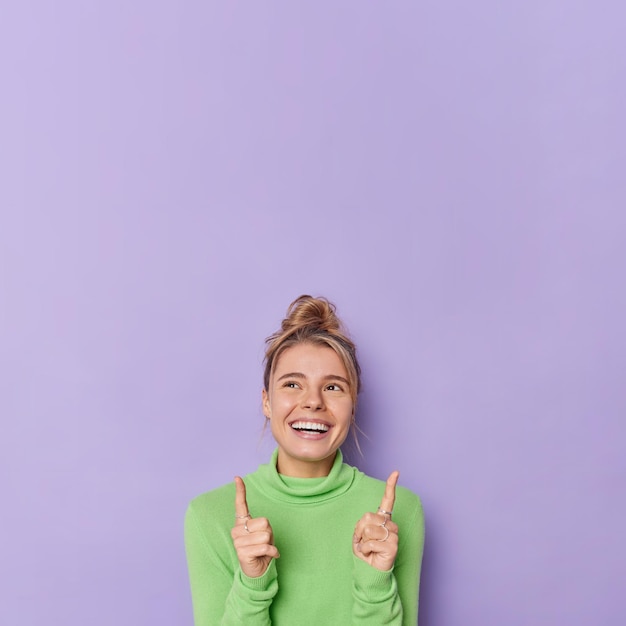 Free photo look what nice offer for you. cheerful optimistic woman points at copy space above suggests to check this demonstrates free place for your advertisement wears green turtleneck isolated on purple wall