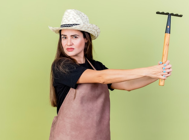 Free photo looking at camera beautiful gardener girl in uniform wearing gardening hat holding out rake at side isolated on olive green background