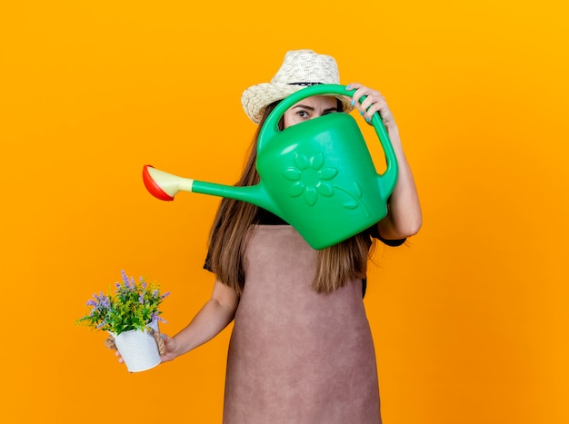 Free photo looking at camera beautiful gardener girl wearing uniform and gardening hat holding flower in flowerpot and covered face with watering can isolated on orange background