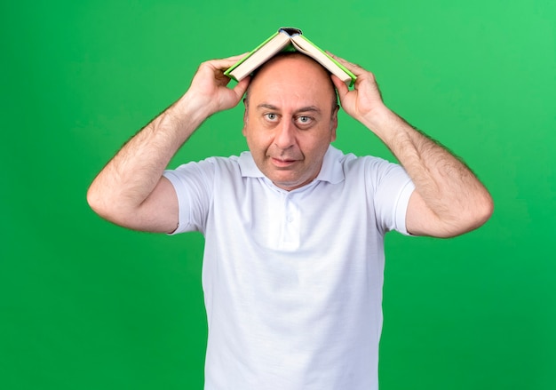 Looking at camera casual mature man covered head with book isolated on green background