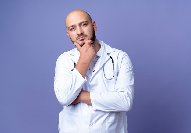 Looking at camera confident young bald male doctor wearing medical robe and stethoscope putting hand on chin isolated on blue background with copy space