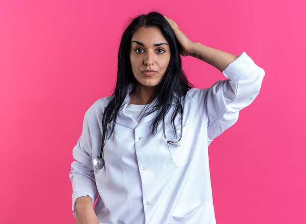 Looking at camera young female doctor wearing medical robe with stethoscope putting hand on head isolated on pink wall