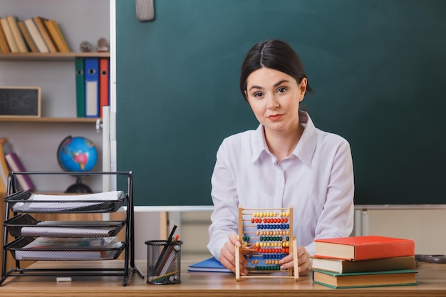 looking at camera young female teacher holding abacus sitting at desk with school tools in classroom