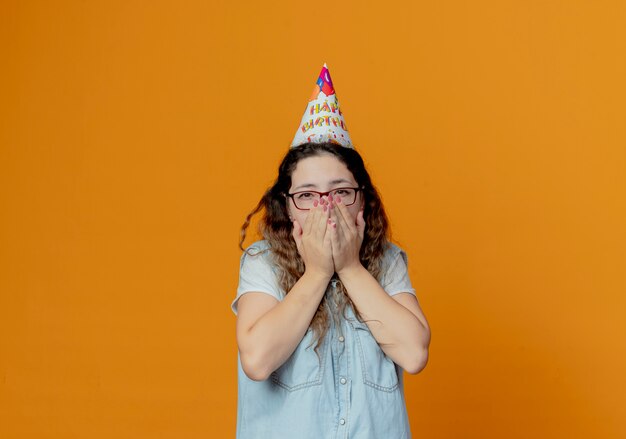 Looking at camera young girl wearing glasses and birthday cap covered with hands mouth isolated on orange background