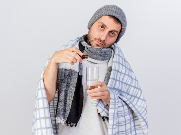 Looking at camera young ill man wearing winter hat and scarf wrapped in plaid pouring medicament