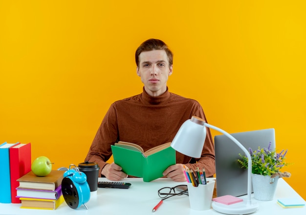 looking at camera young student boy sitting at desk with school tools holding book