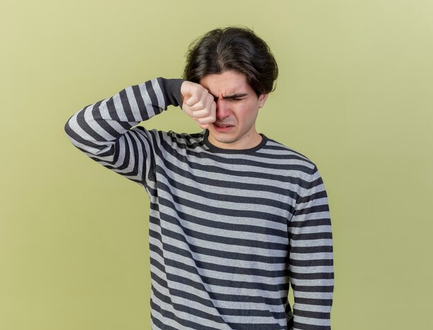 Looking at down crying young ill man wiping eye with hand isolated on olive green background