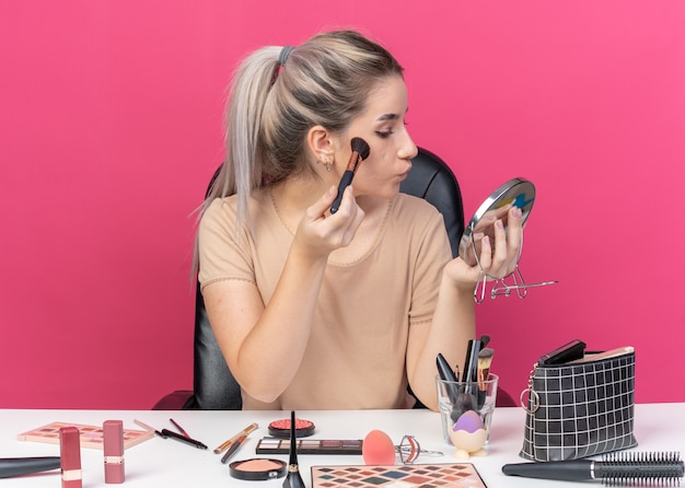 Free photo looking at mirror young beautiful girl sits at table with makeup tools applying powder blush isolated on pink wall