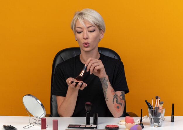 Looking at mirror young beautiful girl sits at table with makeup tools applying powder blush with powder brush isolated on orange wall