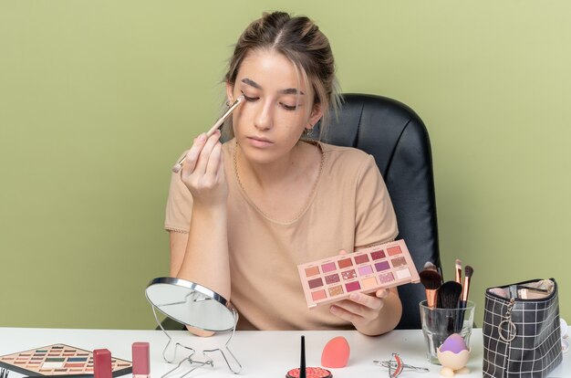 Looking at mirror young beautiful girl sitting at desk with makeup tools applying eyeshadow with makeup brush isolated on olive green wall