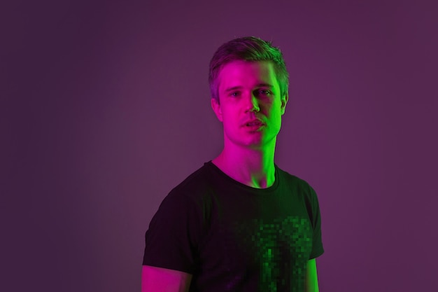 Looking on, serious. Caucasian man's portrait isolated on purple studio background in neon light. Beautiful male model in black shirt. Concept of human emotions, facial expression, sales, ad.