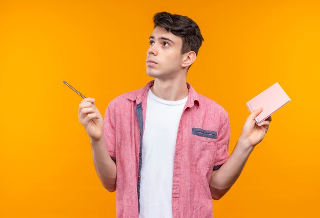 Looking at side caucasian young man wearing pink shirt holding pen and notebook on isolated orange wall