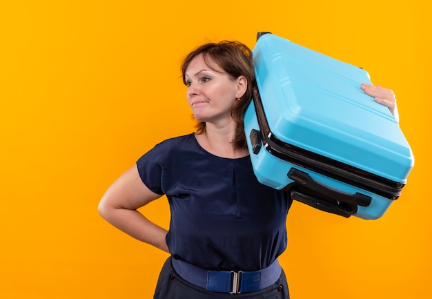 Looking at side confused middle-aged traveler woman holding suitcase on shoulder on isolated yellow wall