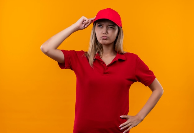 Looking at side confused young delivery girl wearing red uniform and cap putting finger to head isolated on orange background