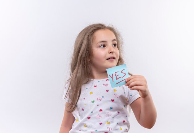 Looking at side little school girl wearing white t-shirt holding paper mark on isolated white wall