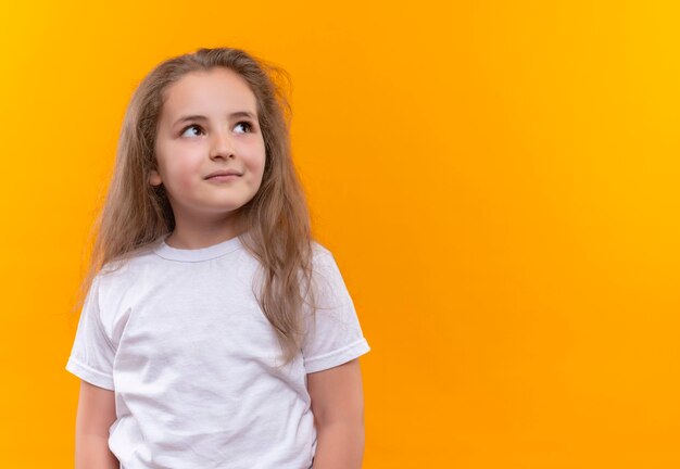 Looking at side little school girl wearing white t-shirt on isolated orange wall