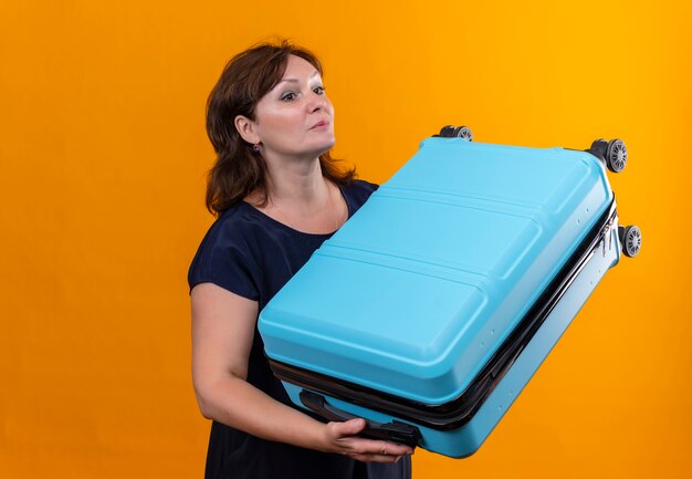 Looking at side middle-aged traveler woman holding suitcase on isolated yellow wall