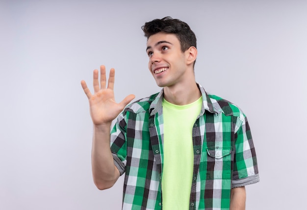 Looking at side smiling caucasian young man wearing green shirt showing five with hand on isolated white wall