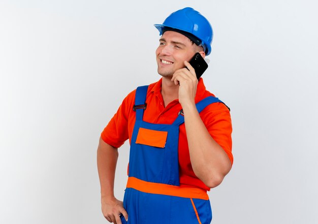 Looking at side smiling young male builder wearing uniform and safety helmet speaks on phone and putting hand on hip 