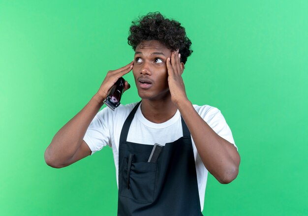 Looking at side surprised young afro-american male barber wearing uniform holding hair clippers and putting hands on forehead