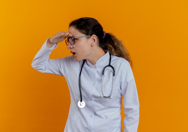 Looking at side surprised young female doctor wearing medical robe and stethoscope with glasses looking at distance with hand isolated on orange wall