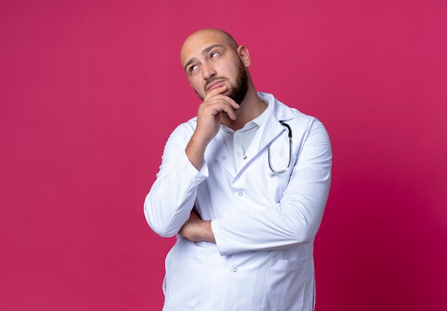 Looking at side thinking young bald male doctor wearing medical robe and stethoscope putting hand on chin isolated on pink background with copy space